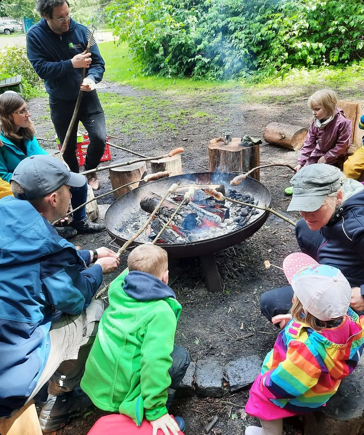 Eine Gruppe Erwachsener und Kinder grillen Würstchen am Sttock über einem offenen Feuer in einer Metallschale | © Sebastian Grumbrecht