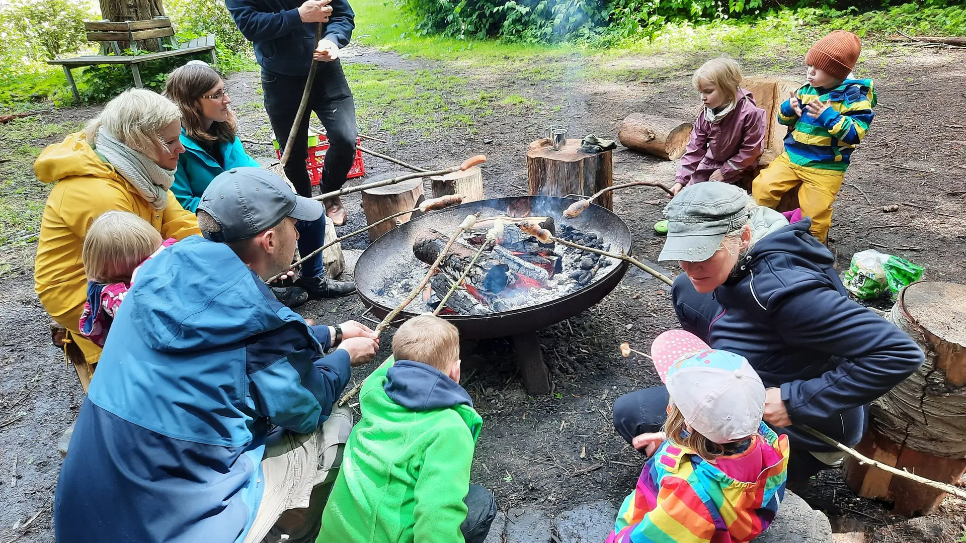 Eine Gruppe Erwachsener und Kinder grillen Würstchen am Sttock über einem offenen Feuer in einer Metallschale | © Sebastian Grumbrecht