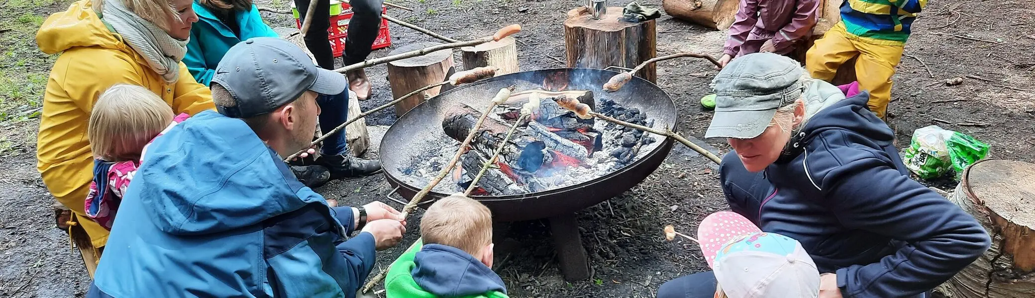 Eine Gruppe Erwachsener und Kinder grillen Würstchen am Sttock über einem offenen Feuer in einer Metallschale | © Sebastian Grumbrecht